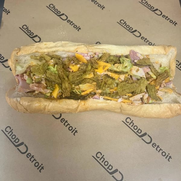 A Chop Detroit sandwich, filled with meat, cheese, lettuce, and crispy green toppings, on branded paper.
