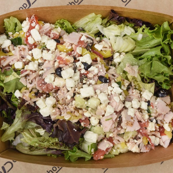 A close-up of a fresh salad in a brown paper bowl, topped with feta, ham, and vegetables.