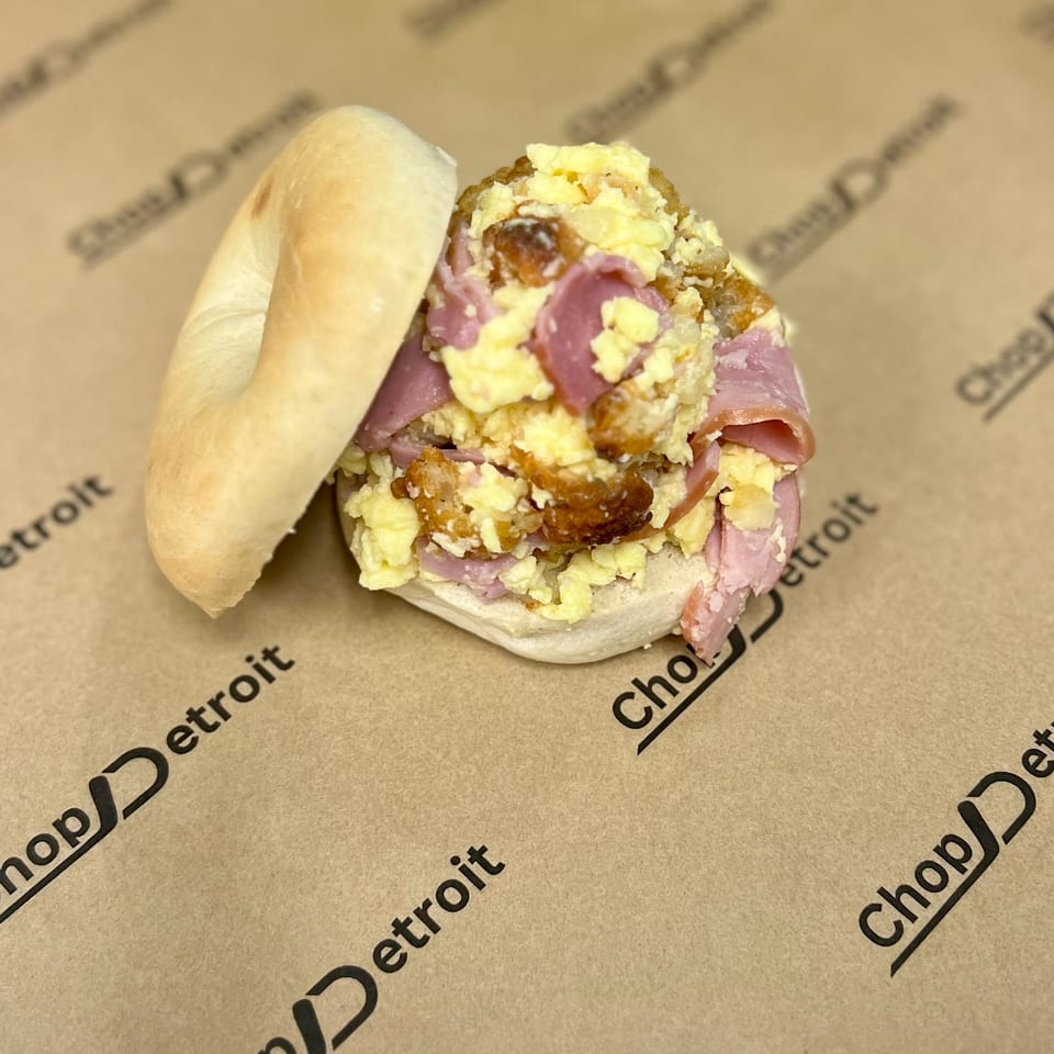 A bagel sandwich from Chop Detroit with scrambled eggs, ham, and crispy bits on branded paper.