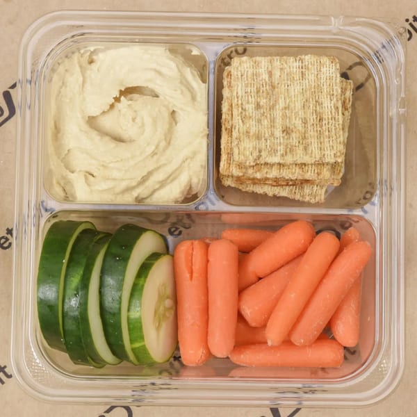 A clear plastic container with hummus, crackers, sliced cucumbers, and baby carrots.